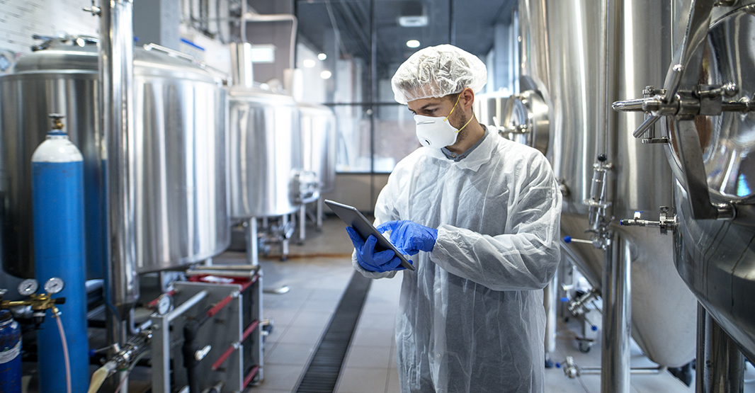 Technologist in white protective uniform controlling industrial process using tablet computer.